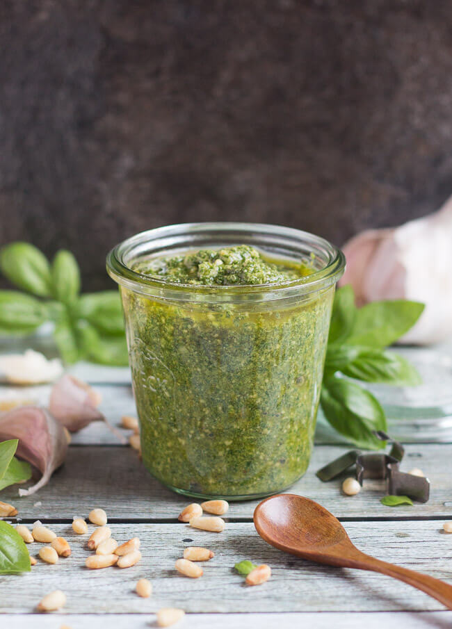 A glass jar of freshly made basil pesto sauce, sitting on a wooden table and surrounded by pesto ingredients. 
