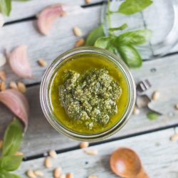 An overhead shot of a jar of basil pesto sauce, surrounded by pine nuts, fresh basil and garlic cloves.