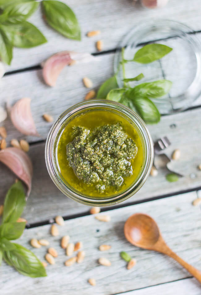 An overhead shot of a jar of basil pesto sauce, surrounded by pine nuts, fresh basil and garlic cloves.