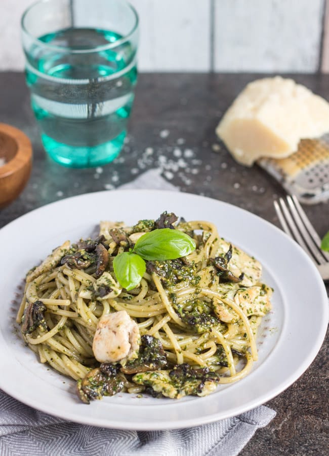 A grey plate piled with chicken pesto pasta.  A blue glass and a chunk of Parmesan cheese sit in the background.  