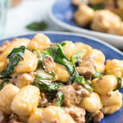 A shot of chorizo & creamy spinach easy gnocchi recipe on a blue plate, with a green water glass in the background.