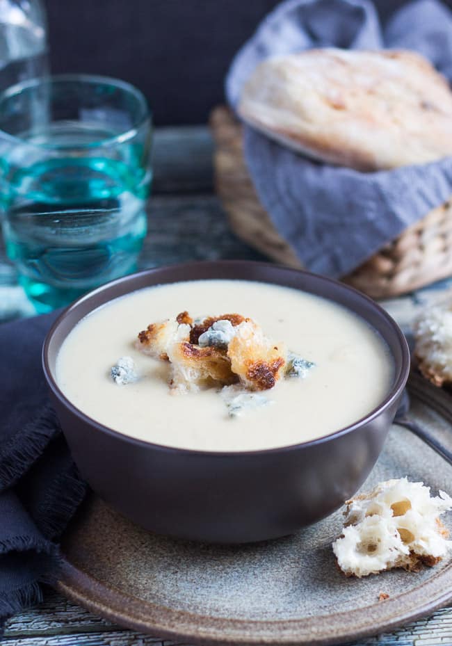 A brown bowl filled with creamy cauliflower cheese soup. Croutons and blue cheese crumbles sit on the top of the soup. 