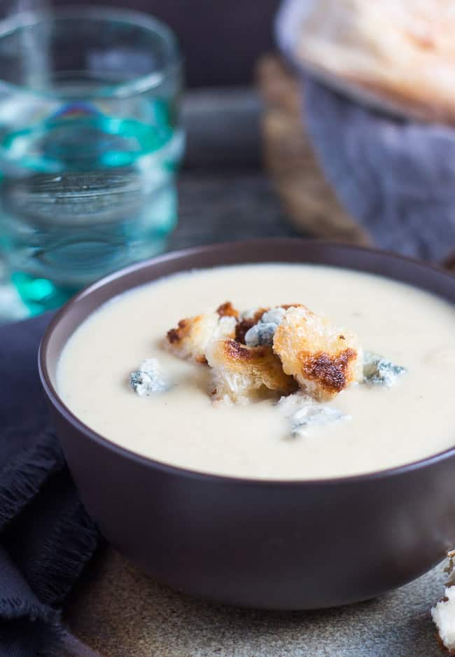 A brown bowl containing this easy cauliflower cheese soup recipe. A blue glass and a loaf of bread sit in the background, behind the soup bowl. 