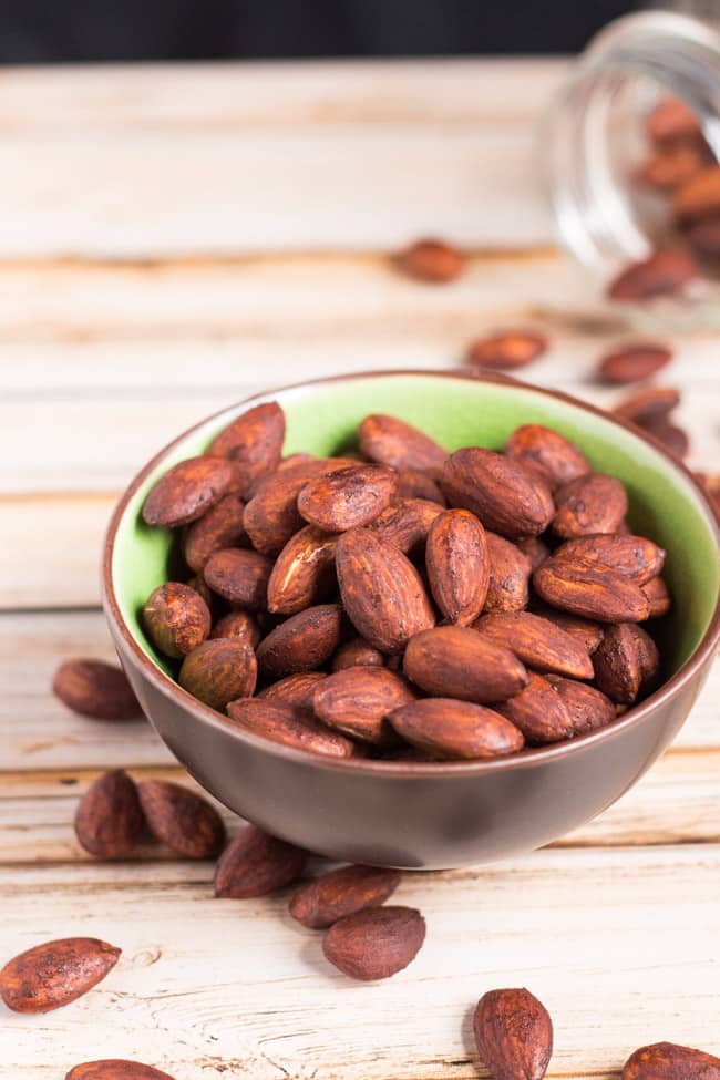 A green and brown bowl full of roasted tamari almonds.  Extra tamari almonds are scattered around the bowl.  