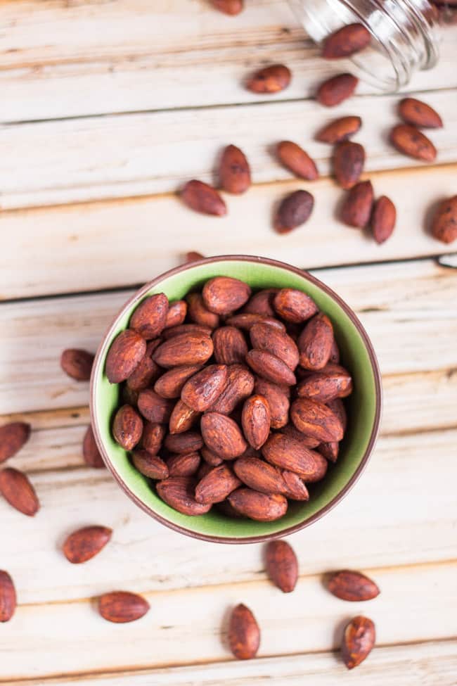 An overhead shot of a green and brown bowl full of roasted tamari almonds. A glass jar at the top is on it's side with almonds spilling out of it.  