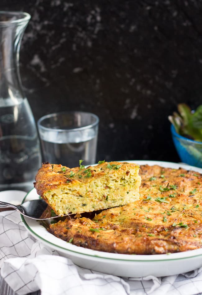 A slice of healthy crustless zucchini quiche balanced on a silver serving spatula, which hovers over the rest of the quiche.  