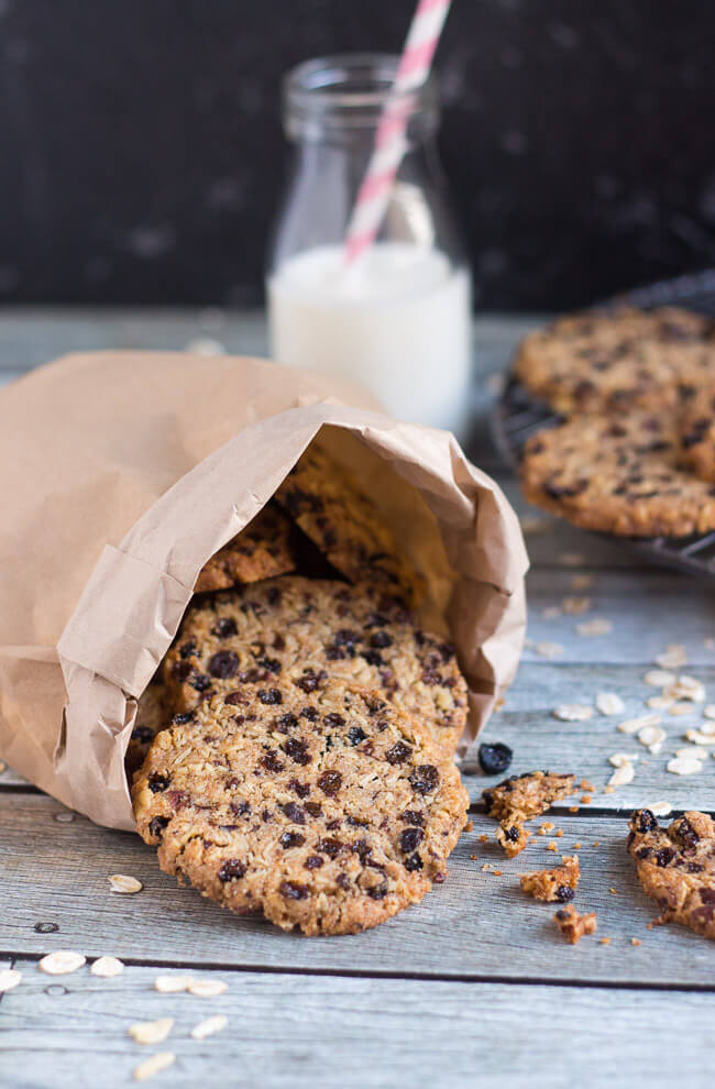 Currant & cocoa nib wholegrain cookies in a brown paper bag, with a bottle of milk and more cookies on a cooling rack. 