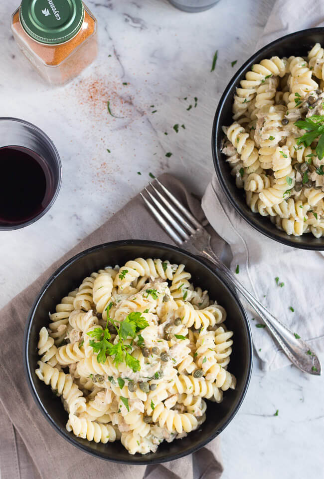 An overhead shot of two black bowls, filled with creamy tuna pasta.  A silver fork, a glass of red wine & a jar of cayenne pepper are dotted around the pasta bowls.  