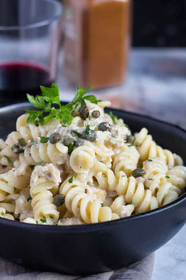 A close up shot of a bowl of creamy tuna pasta, showing the pieces of tuna and capers in the recipe.