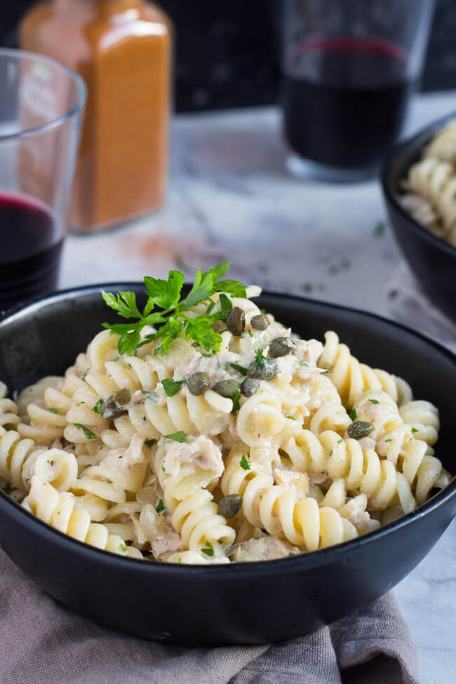 A bowl of creamy tuna pasta, with a large sprig of parsley on top of the pasta, and capers sprinkled over the top.  