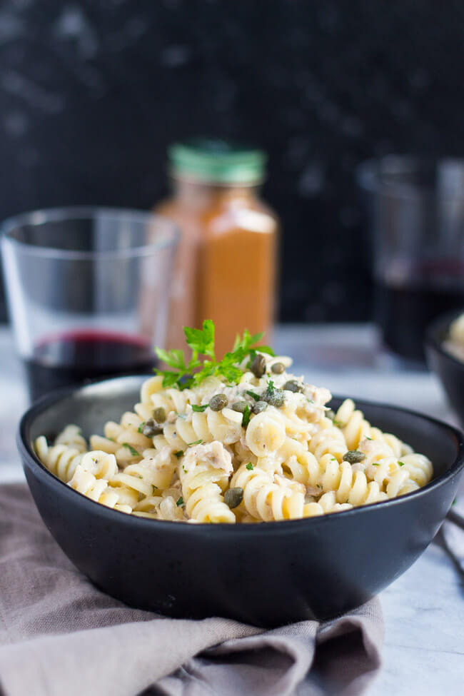 A black bowl full of creamy tuna pasta, with a glass of red wine and jar of cayenne pepper in the background.  