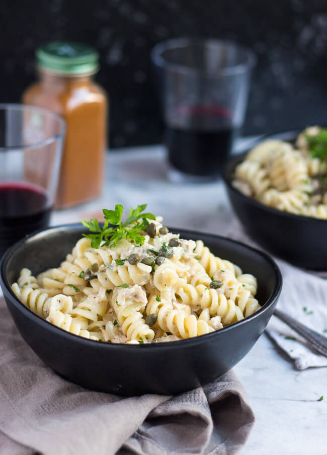 A black bowl, filled with creamy tuna pasta, sitting on a grey napkin.  A second black bowl of creamy tuna pasta sits in the background.  