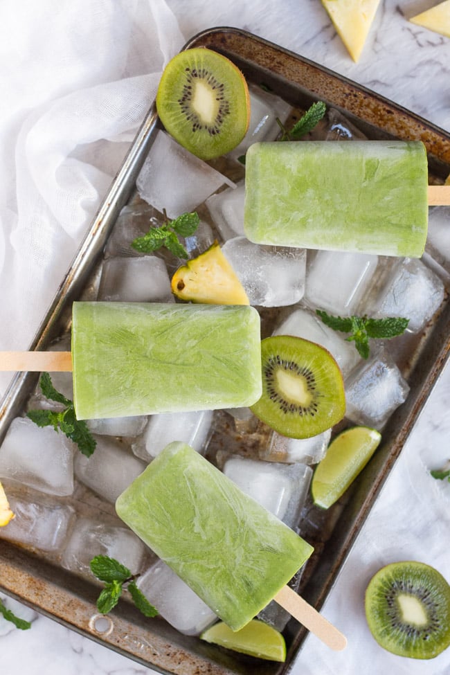 An overhead shot of three green smoothie popsicles lying on a baking tray filled with ice, surrounded by slices of pineaple and kiwi fruit, with sprigs of mint. 