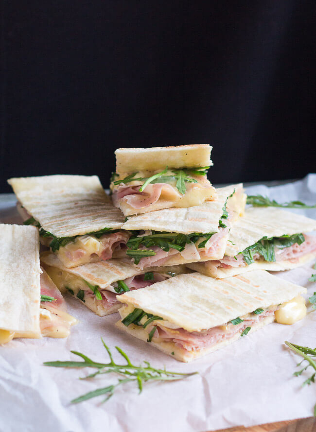 A pile of ham & brie flatbread sandwiches on white paper, surrounded by scattered rocket leaves.