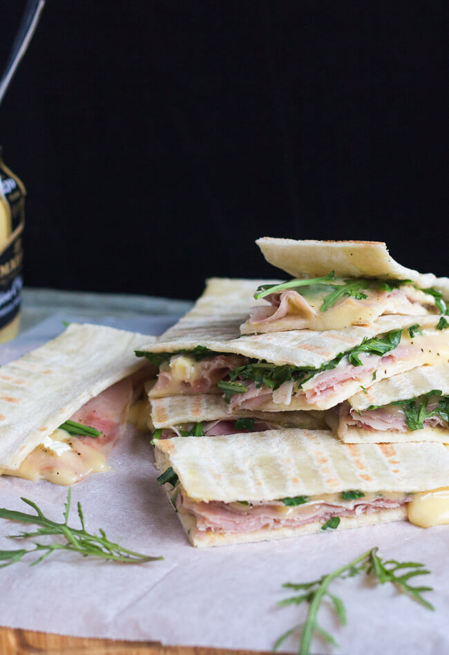 A pile of ham & cheese flatbread sandwiches, with a jar of mustard in the background. 