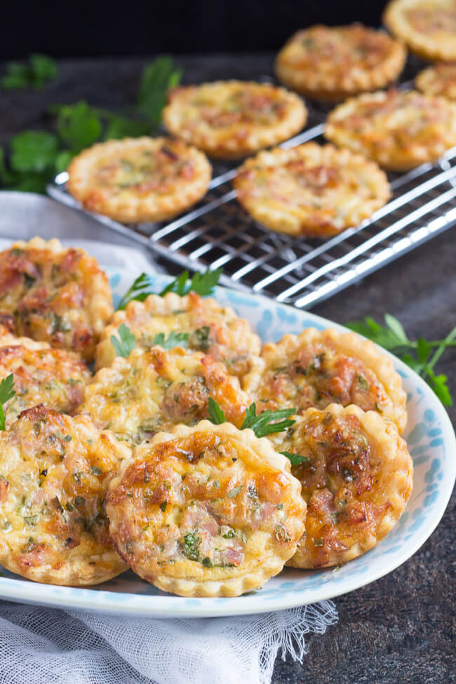 A blue & white platter, piled high with cooked puff pastry mini quiches.  Additional mini quiches are on a cooling rack in the background.