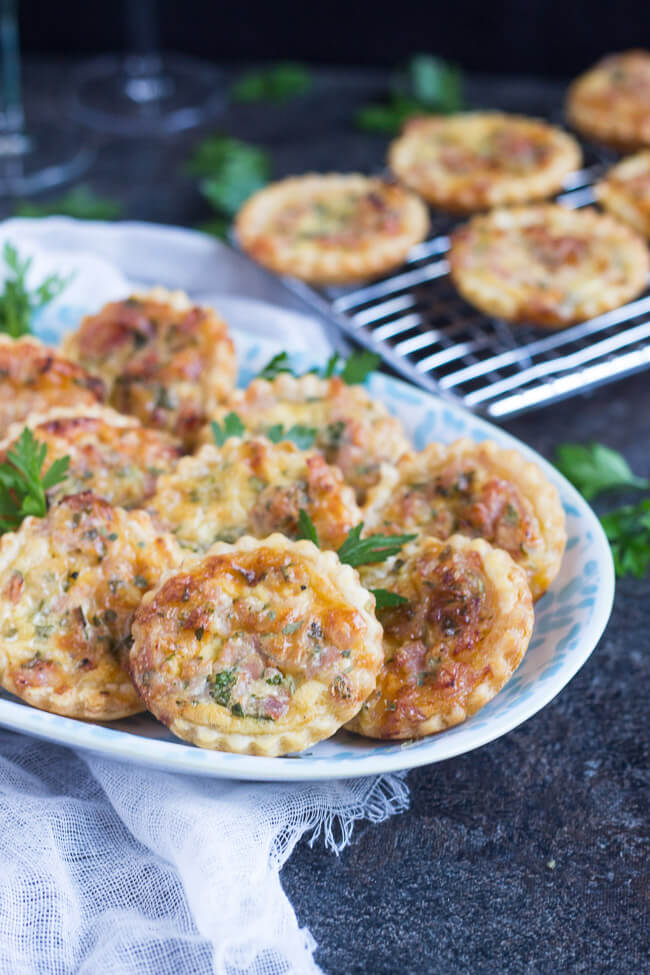 A blue & white platter filled with cooked puff pastry ham and cheese mini quiches that are scattered with parsley.  