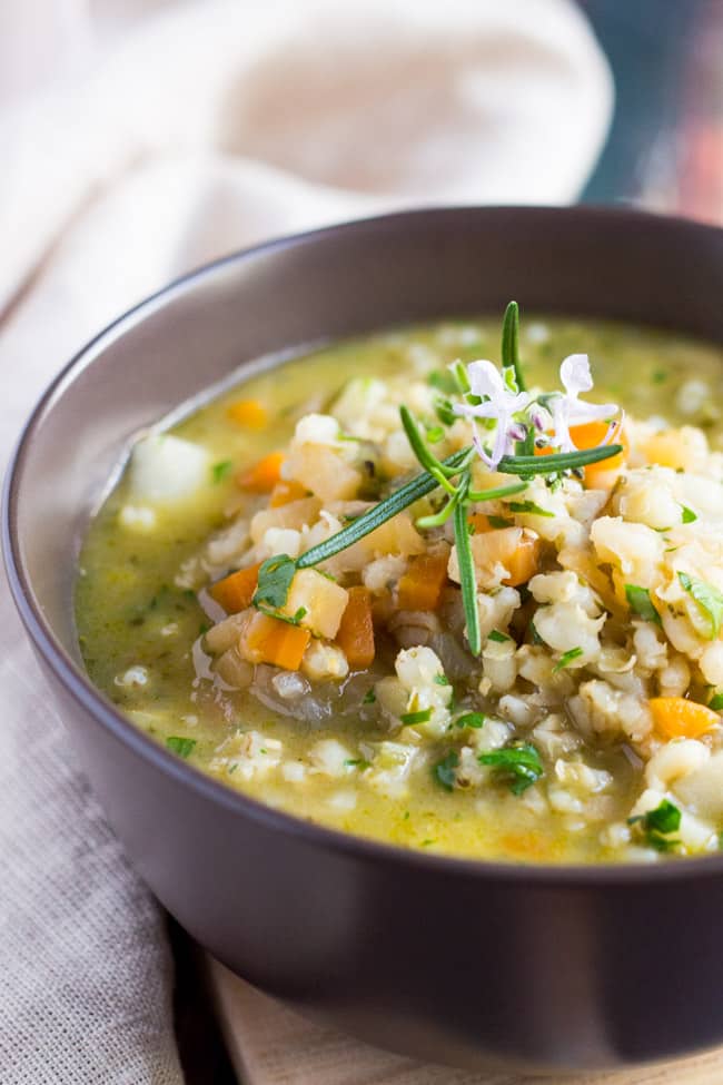 Close up shot of a brown bowl full of Hearty Winter Vegetable Soup. Small pieces of vegetable and swollen grains of barley can be seen in the broth. 