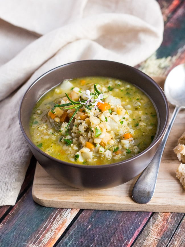 Side shot of a bowl of Hearty Winter Vegetable Soup sitting on a wooden chopping board. A linen napkin sits behind the bowl. . 