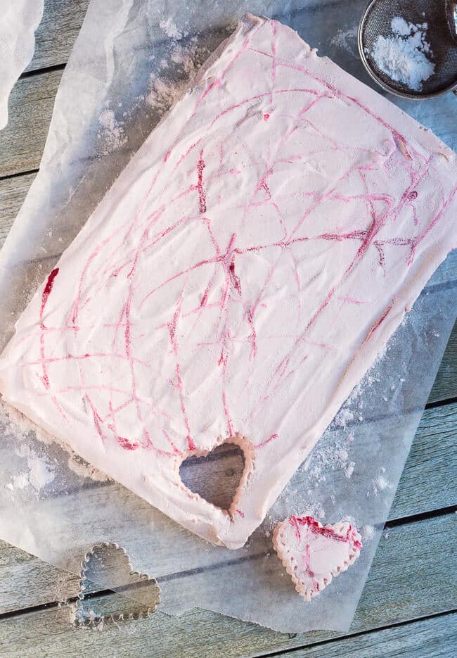 A slab of homemade strawberry marshmallow on a sheet of baking paper, with a single heart shape cut out of the lower right hand corner.