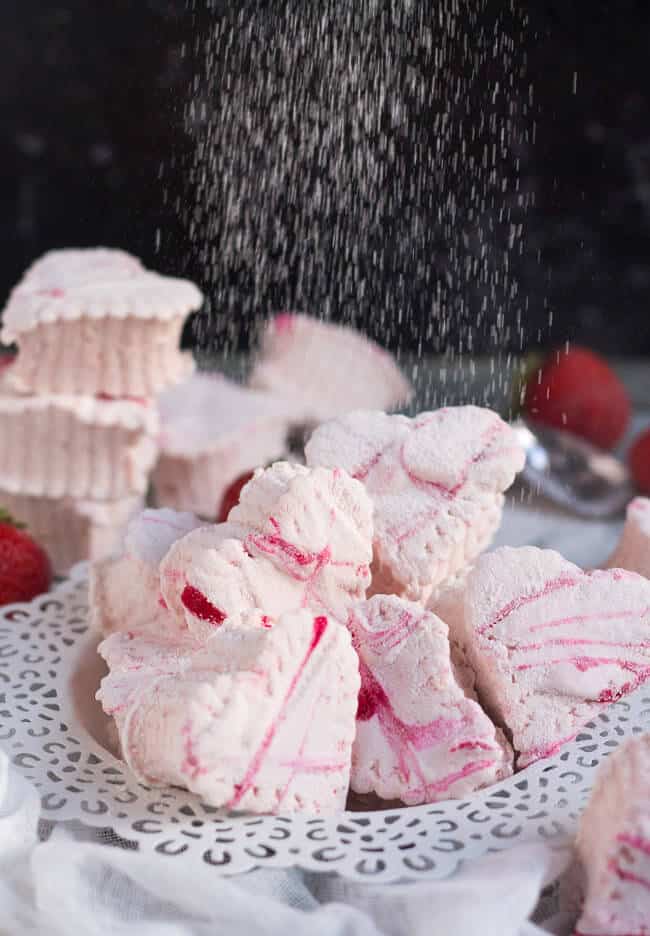 Icing sugar being sifted over a a pile of homemade strawberry marshmallows on a white plate.