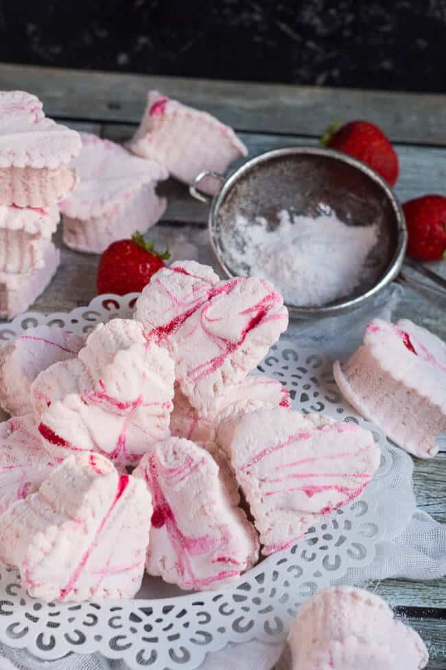 A pile of homemade strawberry marshmallows on a white plate, surrounded by scattered marshmallows and fresh strawberries. 
