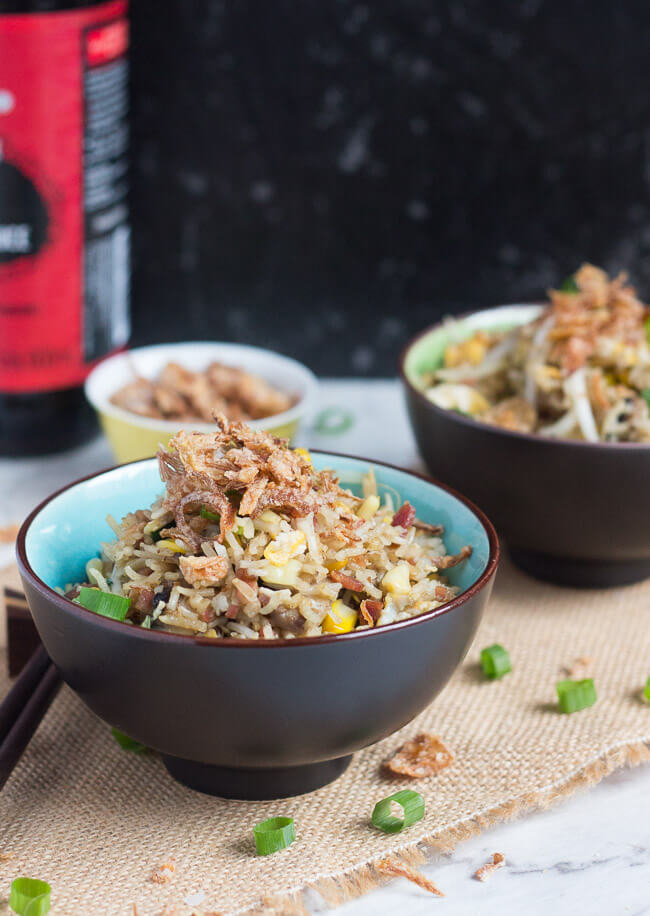 A blue and brown bowl, full of fried rice, with a second bowl of fried rice and a bottle of soy sauce in the background.
