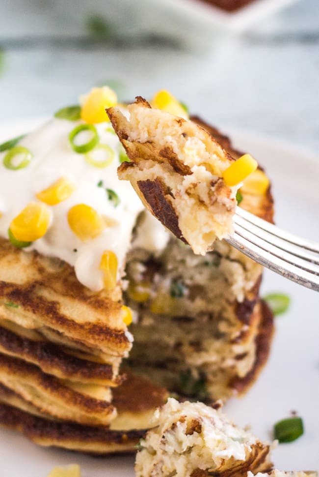 A piece of leftover Mashed Potato & Corn Pancakes on a silver fork, showing the fluffy interior of the pancakes.  The stack of cut pancakes are in the background.