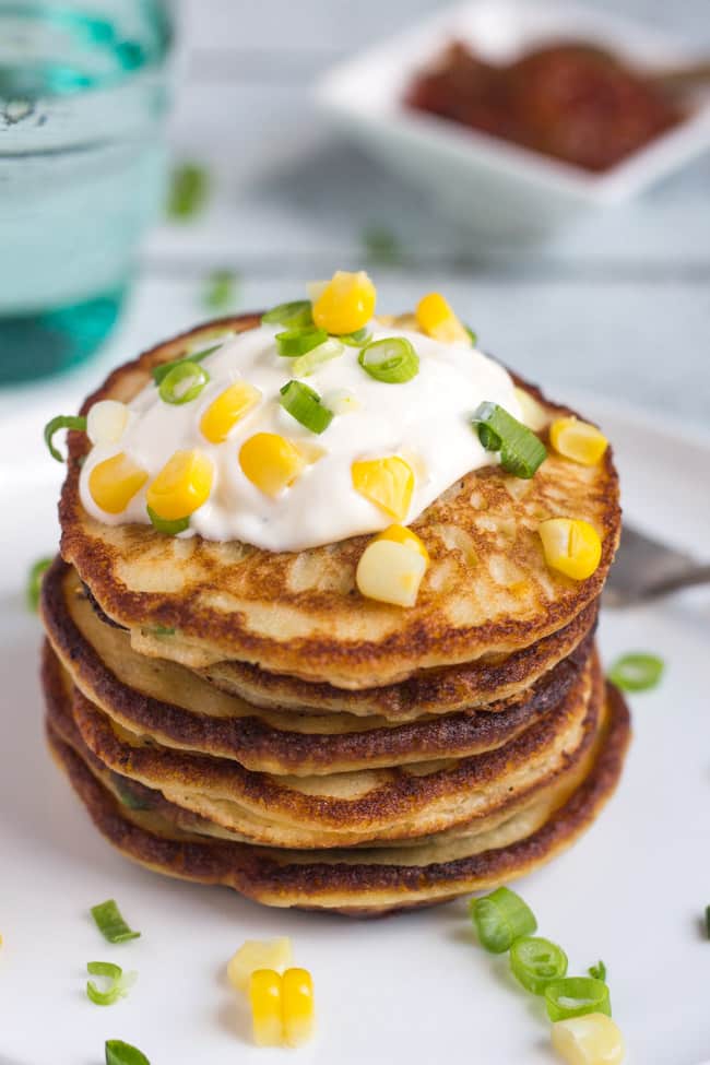 A close up shot of a stack of Leftover Mashed Potato & Corn Pancakes, with fresh corn kernels and chopped spring onions scattered over a dollop of sour cream on top of the pancakes.