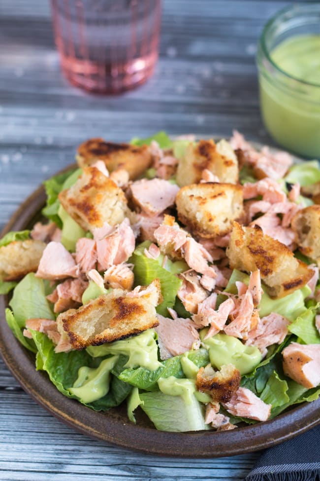 Flaked leftover salmon, croutons, chopped Romaine lettuce, and creamy avocado dressing on a brown earthenware plate with a jar of avocado dressing in the background.  