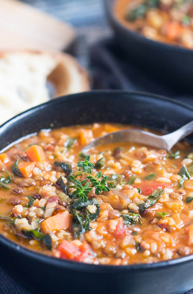 A black bowl, full of lentil and brown rice soup recipe, with chunks of chopped carrot, diced tomatoes and a sprig of thyme on the surface of the soup. 