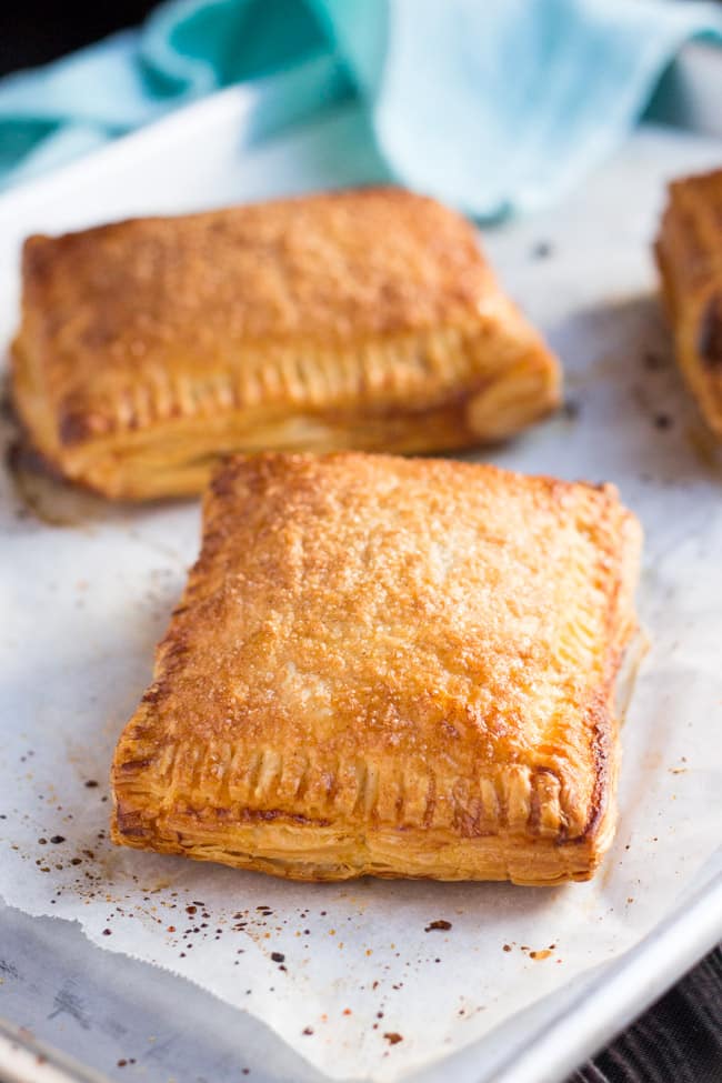 A close up shot of a cooked apple turnover, with two other apple turnovers and a blue tea towel in the background.