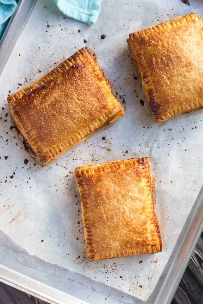 An overhead shot of three cooked apple turnovers, made from an easy apple turnovers recipe, on a lined baking tray with a blue teatowel in the top left hand corner.