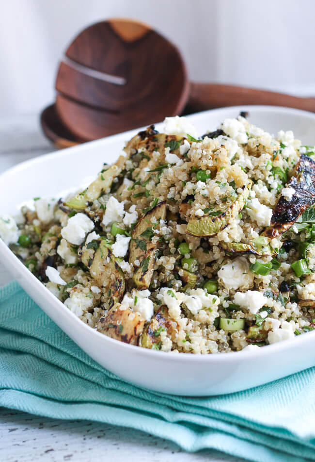 A white plate piled high with quinoa, feta & grilled zucchini salad.  Wooden salad servers sit behind the plate.