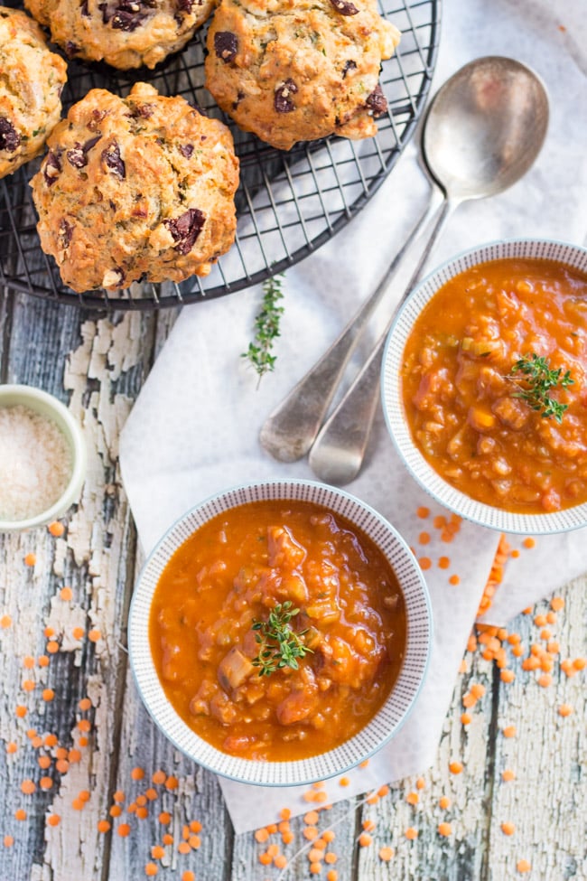 An overhead shot of two bowls of Red Lentil & Smoky Bacon Soup, with a stack of savoury muffins on one side.. 