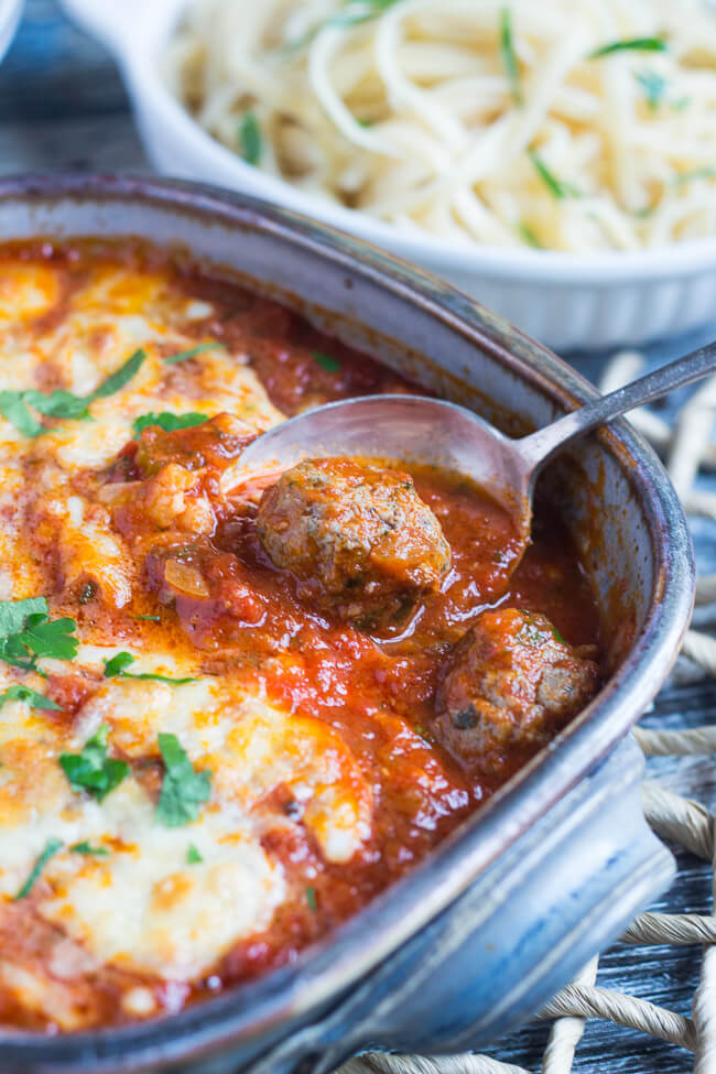Baked Italian Meatballs being spooned out of a blue pottery dish, with a dish of pasta in the background. 