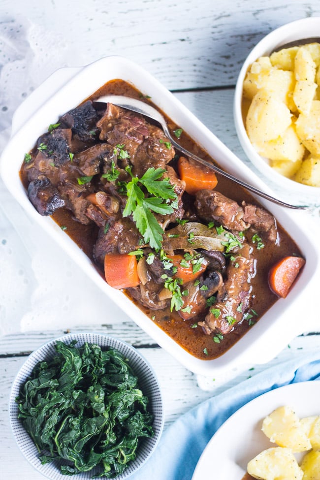 An overhead shot of a white baking dish full of slow cooker beef and mushroom stew, with a large bowl of steamed potatoes, and a bowl of wilted spinach. 