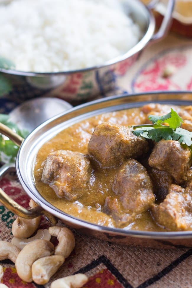 A close up shot of chunks of lamb in a slow cooker lamb korma curry, with a bowl of white steamed rice in the background.  