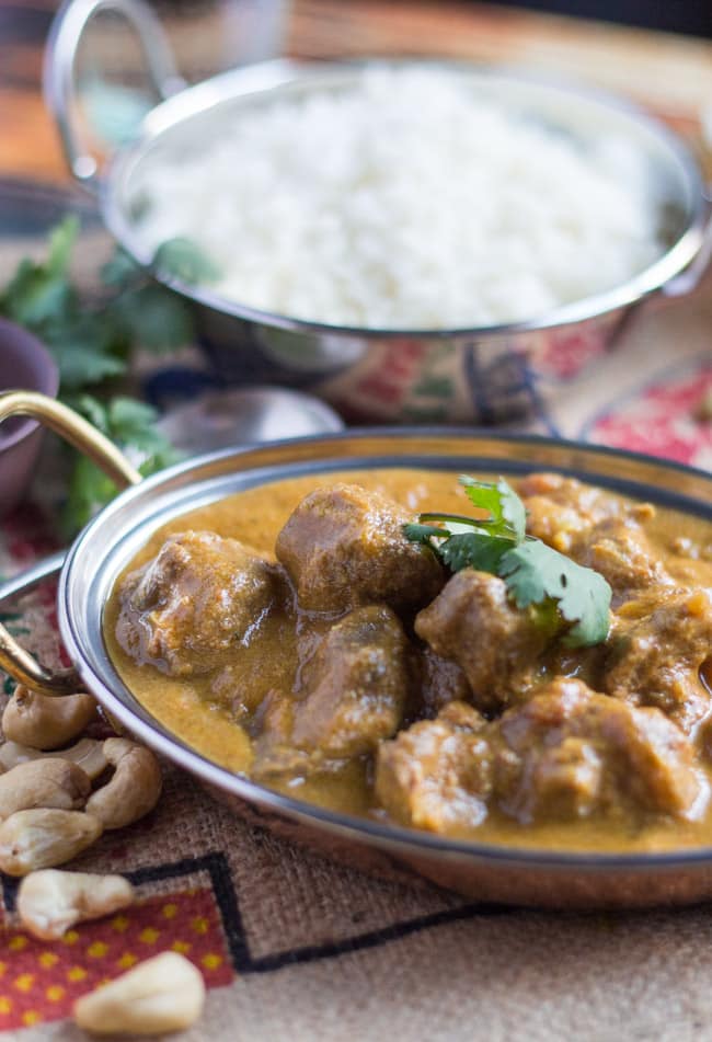 An Indian metal serving dish filled with Slow Cooker Lamb Korma Curry. Another dish of white rice sits in the background of the shot. 