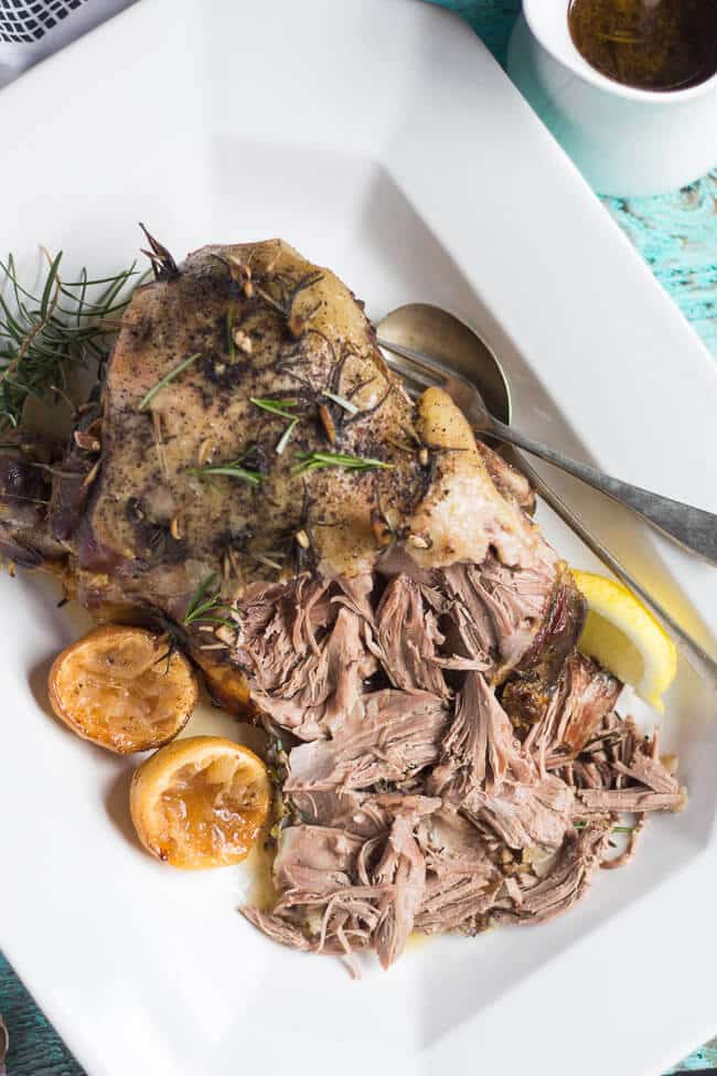 An overhead shot of a cooked slow cooker leg of lamb on a white platter, the bottom edge shredded, surrounded with sprigs of fresh rosemary and lemon halves.  