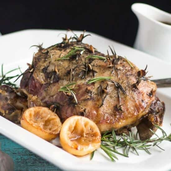 A cooked slow cooker leg of lamb, on a white platter, surrounded by sprigs of fresh rosemary and cooked lemon halves.