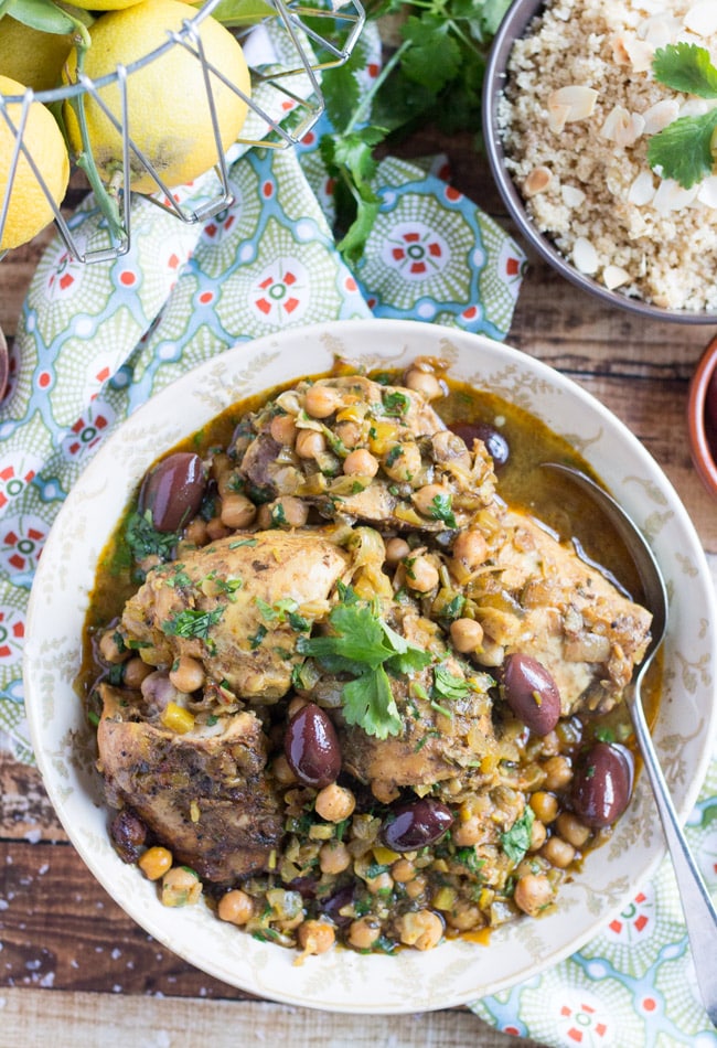 An overhead shot of a white bowl of Slow Cooker Moroccan Chicken Tagine, sitting on a wooden table top, with a bowl of wholemeal cous cous scattered with slivered almonds. .