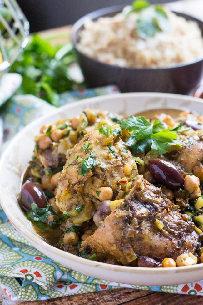 A white bowl holding three pieces of Slow Cooker Moroccan Chicken Tagin, with olives, chickpeas and preserved lemon. A black bowl of wholemeal couscous in the background. 