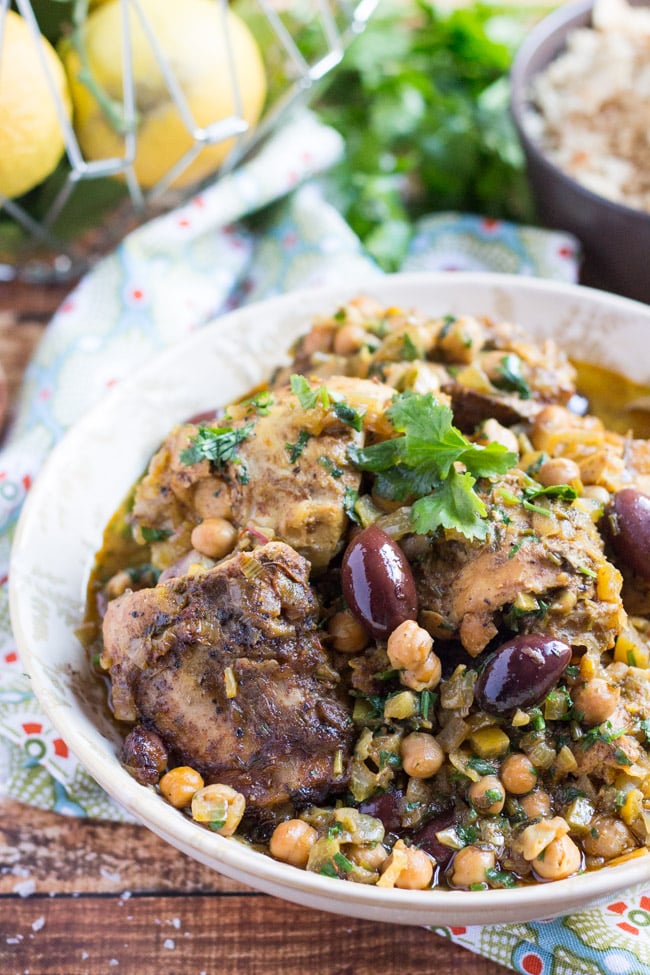 A white bowl full of slow cooker Moroccan chicken tagine, sitting on a blue and green tea towel, with a basket of lemons and a bowl of wholemeal cous cous in the background.