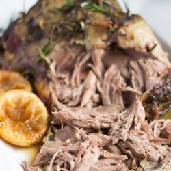 Slow Cooker Lamb Roast sitting on a white platter. The end of the leg has been shredded to display the cooked meat.