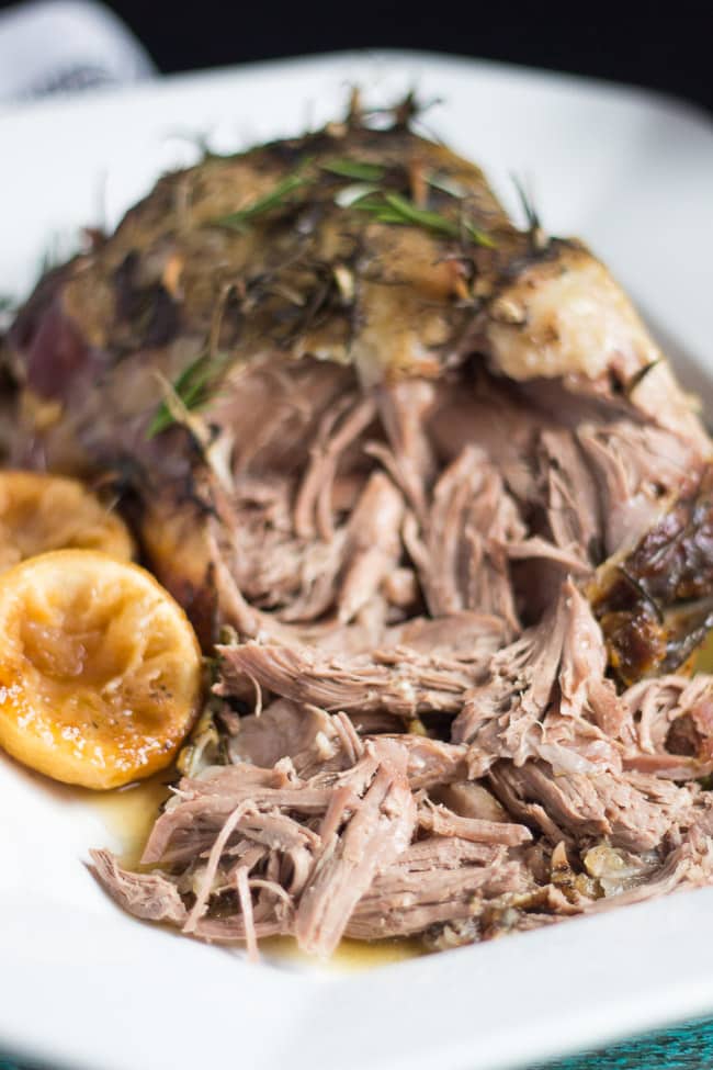 Slow Cooker Lamb Roast sitting on a white platter. The end of the leg has been shredded to display the cooked meat.