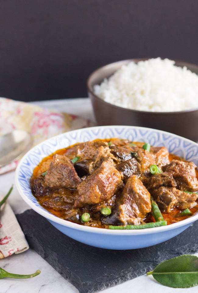 A large blue bowl filled with Slow Cooker Thai Red Beef Curry, with a bowl of steamed rice in the background.