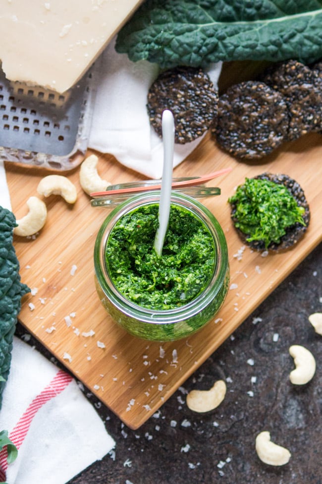 Overhead shot of a jar of spicy kale pesto surrounded by black rice crackers, fresh kale leaves and a chunk of fresh Parmesan cheese.  