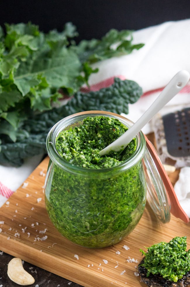 A white spoon inserted in the top of a jar of spicy kale pesto.  A large dollop of spicy kale pesto sits on a black cracker next to the jar.  