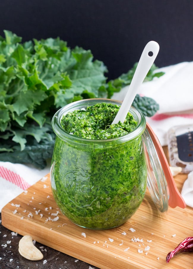 A jar full of spicy kale pesto, with a white spoon in the jar and a bunch of kale in the background.
